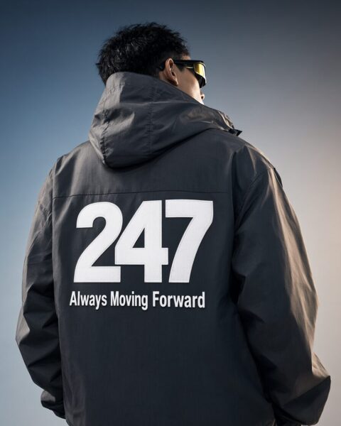 Silhouetted Figure In Monochrome Windbreaker Silhouetted figure wearing dark windbreaker with '247 always moving forward' on back, standing against clear sky.