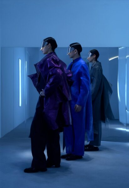 Futuristic Couture In Midnight Hues Three models wearing avant-garde garments in purple, blue, and gray stand in a cool-toned corridor with reflective surfaces.