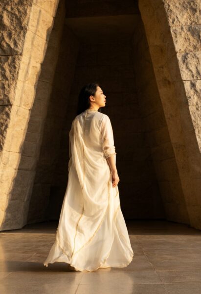 Ethereal White Gown In Angular Light A woman in a flowing white gown with gold embroidery stands within a sunlit stone archway, gazing upward.