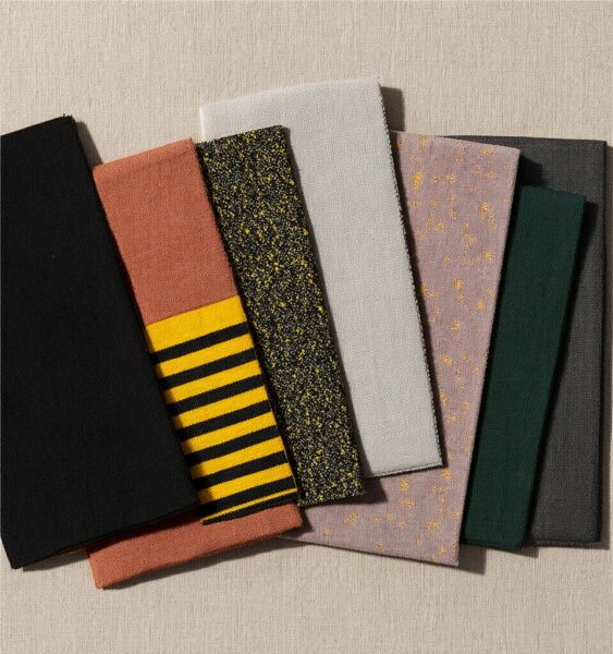 Textured Fabric Swatches In Earth Tones A collection of textured fabric swatches in earth tones including black, terracotta, yellow stripes, speckled gray, mauve, green, and charcoal arranged on a beige surface.
