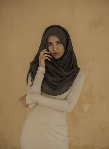 Elegant Veil, Quiet Strength A woman wearing a dark grey hijab and beige long-sleeve dress stands against a textured tan wall, hand near her face.