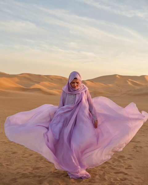 Silken Veil Drifting Through Dunes A woman wearing a flowing lavender hijab and dress stands in a desert, her garments billowing in the wind among golden sand dunes.