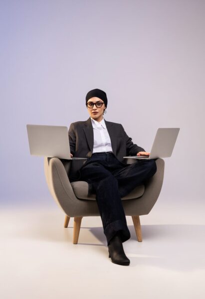 Dual Screen Digital Strategist A woman wearing glasses, a black headscarf, blazer, jeans, and boots sits in a gray chair holding two silver laptops.