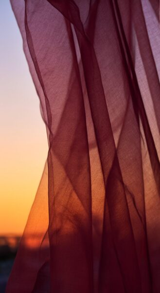 Golden Silk Draped In Sunset Glow Translucent burgundy silk drapes flowing gently against a warm orange and purple sunset sky.