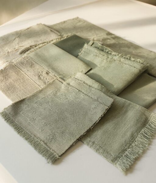 Neutral Textures In Soft Light Soft greenish-gray fabric swatches with frayed edges arranged on a white surface under natural lighting.