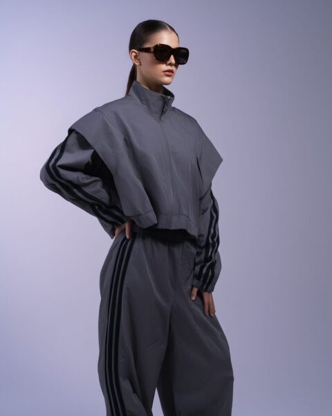 Reflective Silver Tracksuit Reimagined A woman wearing a gray cropped jacket and matching wide-leg pants with three black stripes on each sleeve and leg, posing confidently with sunglasses against a light purple backdrop.