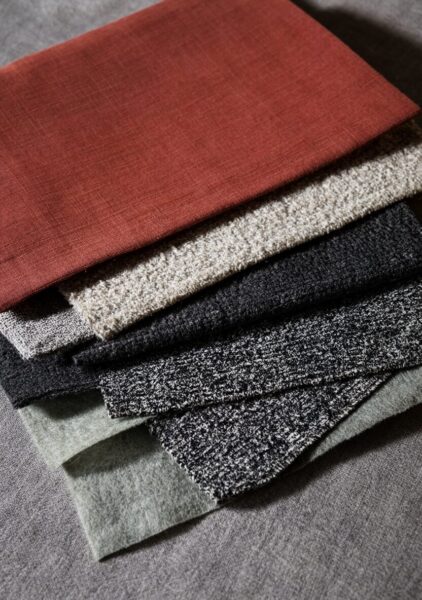 Textured Fabric Swatches In Earth Tones Stacked fabric swatches in earth tones including rust red, beige, black, gray, and green on a textured gray background.