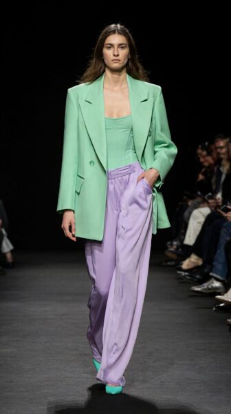 Model Strides In Mint Blazer Ensemble Model walks down runway wearing mint green blazer, lavender wide-leg pants, and matching heels under spotlights.