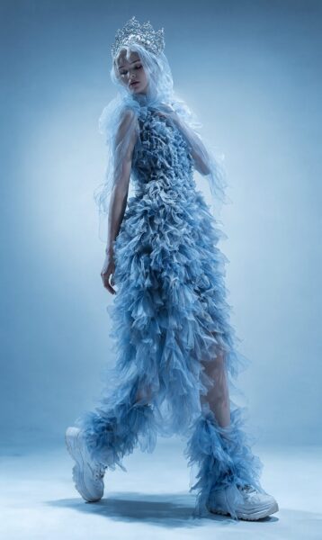 Frost Queen In Azure Ruffles Frost queen in layered azure ruffle dress, wearing silver crown and white sneakers against icy blue backdrop.