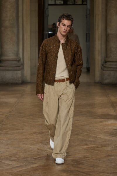 Elegant Brown Ensemble With Gold Accents A male model walks in an elegant brown patterned jacket over a cream knit top, paired with beige wide-leg trousers featuring decorative side trim and white sneakers, set against a grand wooden floor and stone columns.