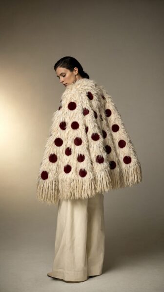 Polka Dot Fur Cape In Soft Ivory A woman wears an ivory fur cape with deep burgundy polka dots, standing in warm studio light against a beige backdrop.