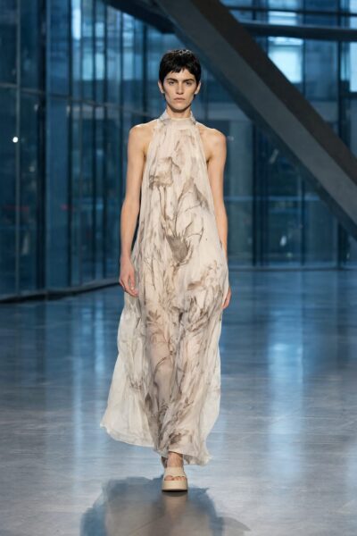 Model Walks In Floral Gown Model walks confidently down a modern glass-walled runway wearing an elegant beige floral-printed halter gown with flowing fabric.