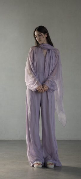 Elegant Mint Jumpsuit With Draped Scarf A woman in a soft lavender jumpsuit with sheer long sleeves and draped scarf stands in minimalist studio space.
