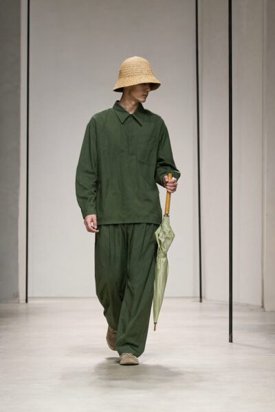 Serene Green Ensemble Walk A model walks down a minimalist runway in an olive green loose-fitting outfit, holding a closed light green umbrella under a woven straw bucket hat.