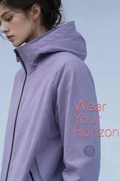 Coral Hoodie Profile Study A woman wears a lavender hooded jacket against a soft blue sky, with the text 'wear your horizon' on her sleeve.