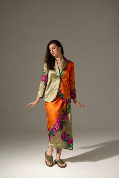 Silk Blazer With Floral Accents A woman wears a silk blazer with floral accents over an orange skirt, standing in soft side-lit studio space.