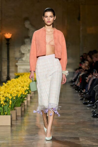 Spring Garden Runway Walk Model walks down a runway lined with yellow tulips wearing a coral cropped jacket, lace bralette, plaid skirt with purple tassels, and light green heels.