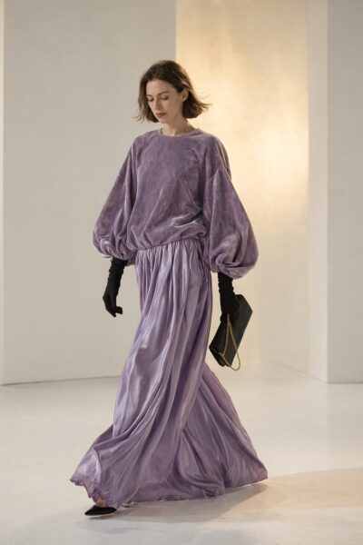 Lavender Velvet Glamour Walk A woman in a lavender velvet ensemble walks gracefully, holding a black clutch with gold chain, under warm ambient light.