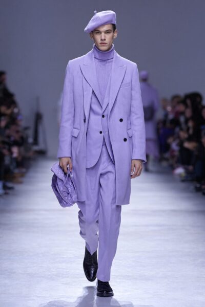 Purple Monochrome Runway Statement A male model walks down a runway in a monochromatic lavender suit, turtleneck, and beret, carrying a matching patterned bag.