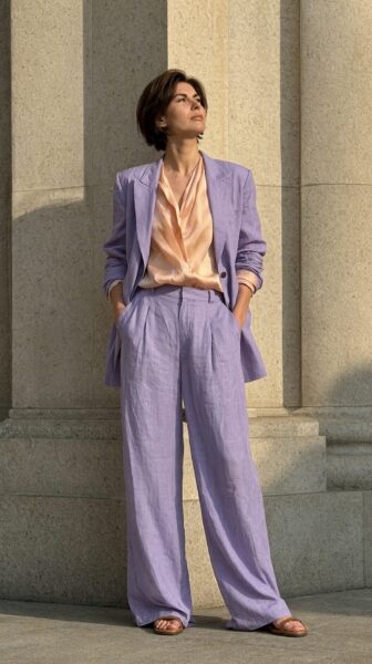 Sunlit Lavender Power Suit A woman in a lavender linen suit stands against stone columns, wearing a peach silk blouse and brown sandals under warm sunlight.