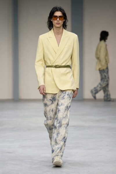 Soft Yellow Suit With Earth-Tone Accents Model walks down runway wearing soft yellow blazer with olive belt and tie-dye pants under neutral lighting.