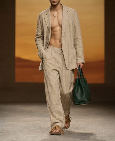 Desert Wind Ensemble Walk A male model walks on a runway wearing an open beige linen suit, fringed sandals, and holding a dark green leather tote bag against a warm-toned desert-inspired backdrop.