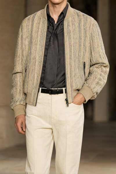 Elegant Earth Tones Ensemble Man wearing beige textured bomber jacket, black shirt, cream trousers, and black belt in elegant indoor setting.
