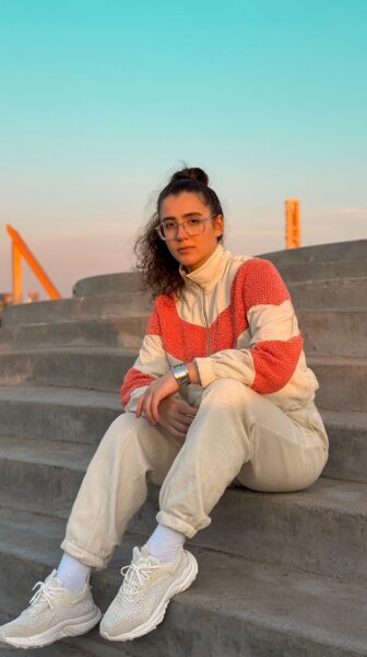 Urban Zen In Coral And Cream A young woman sits on concrete stairs wearing a coral and cream track jacket, beige joggers, white socks, and chunky sneakers under soft sunset light.