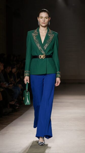 Emerald Blazer With Gold Trim Model walks runway in emerald blazer with gold trim, blue wide-leg pants, holding green purse.