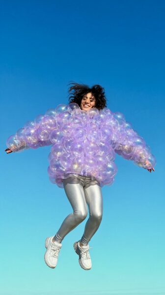 Joyful Leap In Bubble Jacket A person leaps joyfully wearing an iridescent purple bubble jacket against a gradient blue sky.