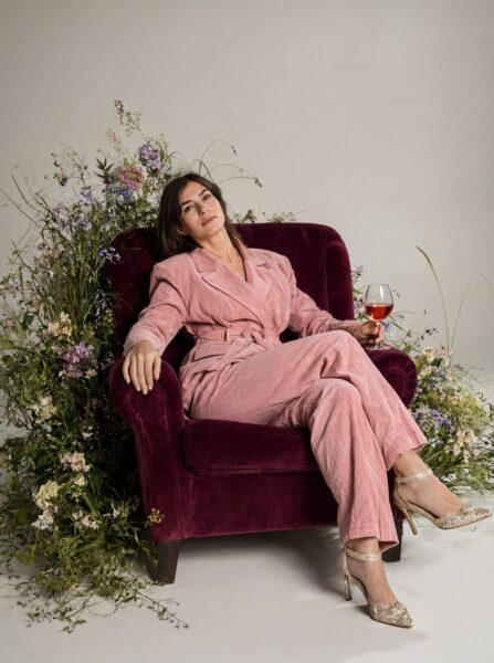Pink Velvet Lounge In Floral Embrace A woman lounges in a plush pink corduroy robe on a velvet chair surrounded by lush wildflowers while holding a glass of rosé wine.