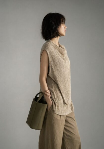 Elegant Asymmetrical Sleeveless Top A woman in profile wears an elegant beige asymmetrical sleeveless top and wide trousers, holding an olive green tote bag against a neutral gray background.
