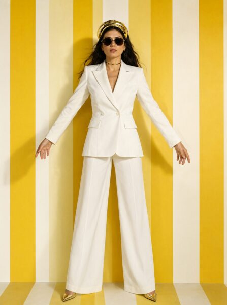Confident White Suit Against Bold Stripes Confident woman in white suit stands against bold yellow and white striped wall wearing sunglasses and gold accessories.