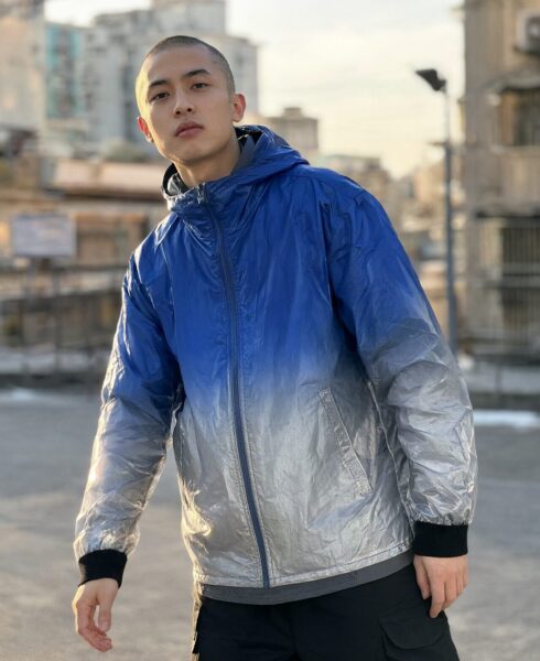Urban Explorer In Gradient Anorak Urban explorer wearing blue-to-silver gradient anorak, standing outdoors on city street during golden hour.