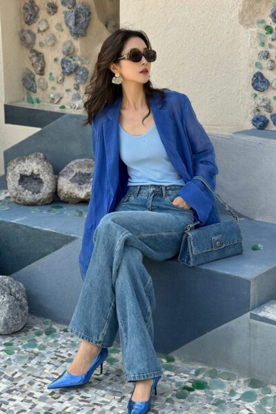 Oceanic Blue Serenity A woman in a blue blazer, light blue top, wide-leg jeans, and metallic blue heels sits on stone steps surrounded by geodes and sea glass mosaic tiles.