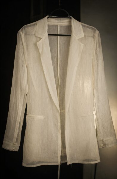 Transparent Blazer With Pearl Accents A sheer white blazer with pearl button accents on cuffs, hanging against dark background.