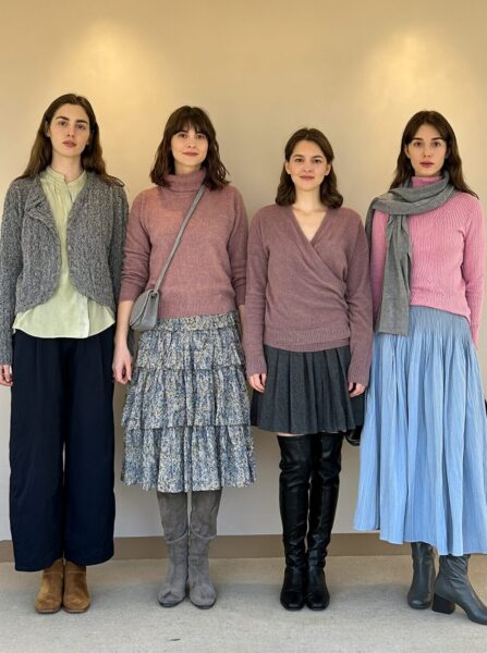 Soft Pastel Winter Ensemble Four women pose in soft pastel winter ensembles featuring sweaters, skirts, boots, and scarves against a warm beige wall.