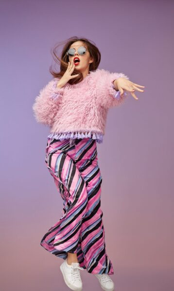Joyful Jump In Fuzzy Pink A woman joyfully jumping mid-air wearing a fluffy pink jacket, colorful striped pants, and white sneakers against a lavender backdrop.