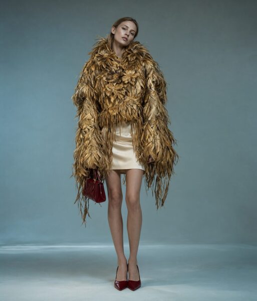 Golden Feathers Embrace A woman in a voluminous golden feathered coat over a cream satin mini dress, holding a red handbag, standing against a muted teal studio background.