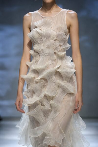Organic Pleated Sculpture Dress A model wears an elegant off-white gown featuring cascading fan-like pleats, showcased on a runway under dramatic stage lighting.