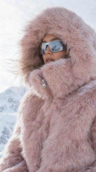 Pink Snow Goddess In Fuzzy Winter Gear A woman wearing a fluffy pink winter coat and silver-framed sunglasses stands in a snowy mountain landscape.