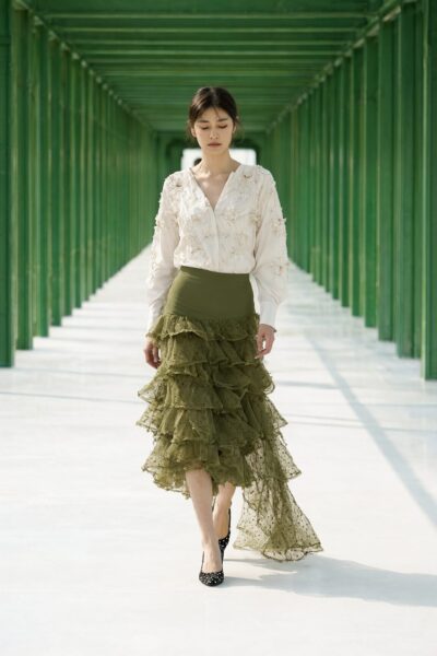 Model Walks Under Geometric Canopy Model walks confidently under a green metal canopy wearing an off-white embroidered blouse and olive green ruffled asymmetrical skirt.