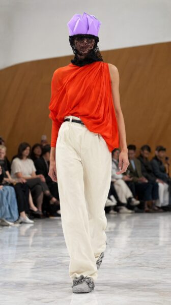 Model Walks Runway In Avant-Garde Ensemble Model walks runway wearing vibrant orange draped top, cream wide-leg pants, and lavender origami-style hat with black lace veil.