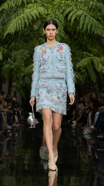Model Walks Through Lush Greenery Model walks confidently down a runway lined with ferns, wearing a light blue floral tweed suit with fringed hems and carrying a silver bird-shaped purse.