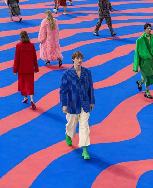 Striding Through Colorful Waves A model strides confidently through vibrant blue and coral waves wearing a blue pinstripe blazer, cream pants, and bright green boots.