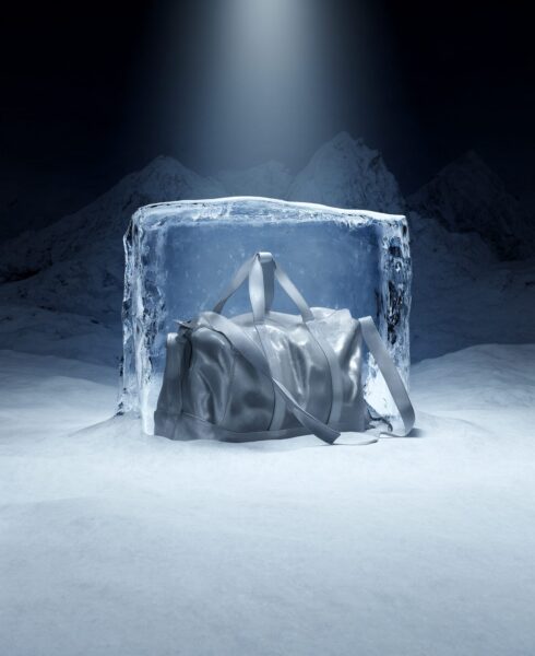 Frozen Duffle Bag In Glacier Ice A silver duffle bag encased in a large transparent ice block, set against snowy mountains under cool blue lighting.