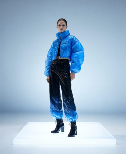 Model In Futuristic Blue Puffer Outfit Model wears futuristic cropped blue puffer jacket and glossy black balloon pants on illuminated white platform against light blue backdrop.