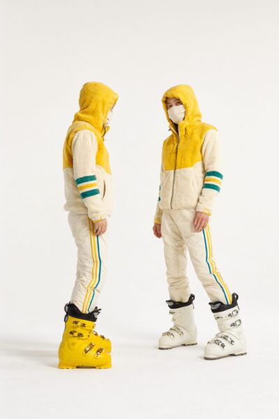 Two Models In Vibrant Ski Gear Two models wearing vibrant yellow and cream ski jackets with striped sleeves, paired with matching pants and contrasting ski boots, standing against a white background.