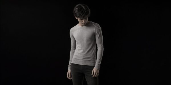 A man in a gray long-sleeve sweater stands alone against a black backdrop, looking down with a silver chain draped over his shoulder.