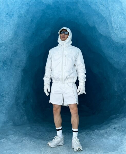 A man stands full-length in a white hooded jacket and shorts inside an icy blue glacier cave wearing white gloves and sneakers.